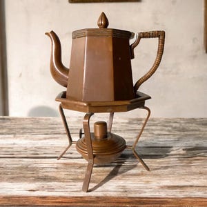 May include: A copper teapot with a wooden handle sits on a copper stand with three legs. The teapot has a hexagonal shape and a pointed lid.