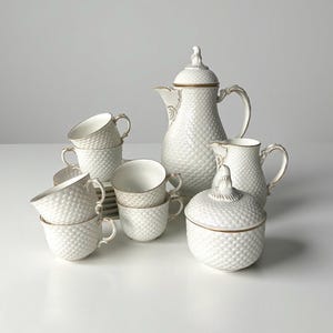 May include: A white porcelain tea set with a textured, quilted pattern. The set includes a coffee pot, creamer, sugar bowl, and several teacups and saucers. Gold trim accents the edges of the pieces. The set is arranged on a white surface.