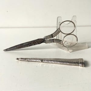 May include: Antique silver sewing scissors with a matching sheath. The scissors have a pointed blade and round finger holes. The sheath is intricately decorated with a repeating pattern. The scissors are displayed on a white surface.