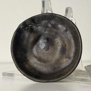 May include: A small, antique bronze bowl with a circular design. The bowl has a dark, aged patina and a small, embossed symbol at the center. The bowl is displayed on a clear stand.