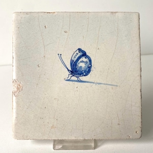 May include: A square ceramic tile with an off-white, crackled glaze finish. Centered on the tile is a hand-painted blue butterfly illustration. The butterfly is depicted in profile with delicate line work and a small shadow cast beneath it.