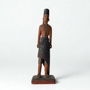 Può includere: Una statuetta in legno intagliato di un uomo con un cappello alto nero, baffi e un drappo scuro. La figura ha un busto e gambe marroni e si trova su una base rettangolare. È un'opera d'arte popolare tradizionale.