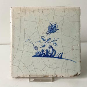 Pode incluir: Azulejo cerâmico quadrado, branco sujo, com um desenho floral azul. O desenho inclui um inseto estilizado, uvas e folhas. O azulejo tem uma aparência rachada e envelhecida, sugerindo que é vintage ou antigo.