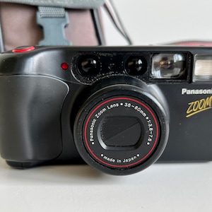 May include: Black Panasonic Zoom camera with a 38-80mm lens. The camera has a red button and a flash. The lens has the text "Panasonic Zoom Lens" and "made in Japan". A gray camera bag is in the background.