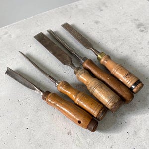 Puede incluir: Un conjunto de cinco cinceles de madera antiguos con hojas de metal y mangos de madera. Los cinceles tienen diferentes formas de hoja, incluyendo bordes planos y curvos. Los mangos son de madera de color marrón claro.