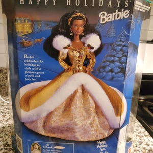 May include: A black Barbie doll wearing a gold gown with white faux fur trim. The doll is holding a red and gold ornament. The box has the text "Happy Holidays Barbie" on it.
