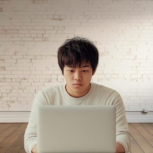 Könnte beinhalten: Eine Person sitzt vor einer weißen Backsteinwand und schaut auf einen Laptop.
