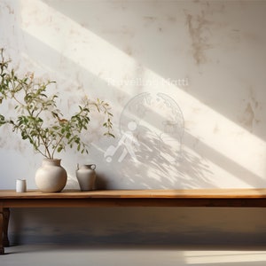 May include: A wooden bench with a light brown finish sits in front of a white wall with a light brown plant in a white ceramic vase. The wall has a light brown textured finish and a shadow from a window.