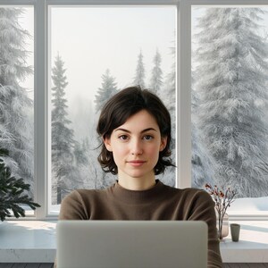 May include: A woman with short brown hair sits in front of a large window with a snowy forest scene. She is looking at the camera and has a laptop in front of her.