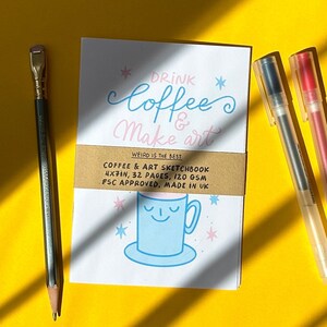 Puede incluir: Un cuaderno de bocetos blanco con el texto "Drink Coffee & Make Art" y una ilustración de taza azul, acompañado de un lápiz y dos bolígrafos. El cuaderno mide 10x18cm y tiene 32 páginas. El fondo es amarillo.