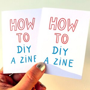 May include: Two white cards with the text "How to DIY a Zine" in red and blue lettering. The cards are held in a hand.