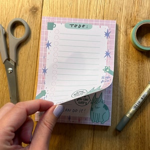 以下が含まれることがあります： ピンク色の格子柄のデザインで、上部に「TODO:」という文字が入ったメモ帳。メモ帳には猫のイラストと「Now you can nap」と「You can do it！」という文字が入っています。ハサミ、ペン、マスキングテープがメモ帳の隣にあります。