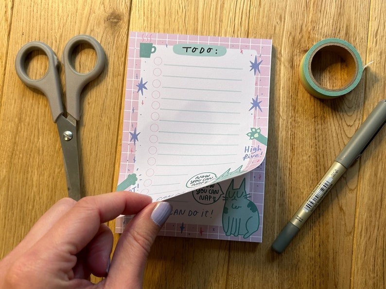 以下が含まれることがあります： ピンク色の格子柄のデザインで、上部に「TODO:」という文字が入ったメモ帳。メモ帳には猫のイラストと「Now you can nap」と「You can do it！」という文字が入っています。ハサミ、ペン、マスキングテープがメモ帳の隣にあります。