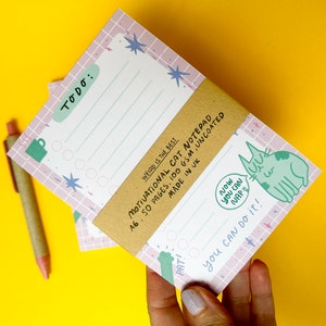 以下が含まれることがあります： ピンクと白の格子柄のA6サイズの「Motivational Cat」ノート。緑色の猫のイラストと「Now you can nap!」と「You can do it!」の文字が特徴です。「To Do:」セクションが含まれています。50ページ、100 gsm、無コーティング。