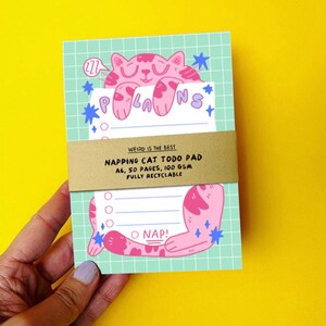 K&ouml;nnte beinhalten: Notizblock mit einer rosa Katzenillustration, dem Wort "PLANS" und dem Text "NAPPING CAT TODO PAD". Der Block hat das Format A6, 50 Seiten und 100 g/m&sup2; Papier. Der Notizblock wird von einer Person gehalten.