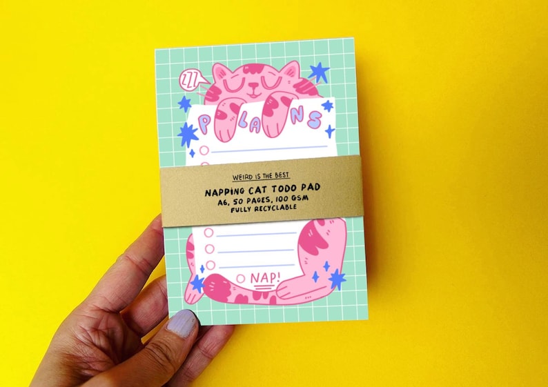 K&ouml;nnte beinhalten: Notizblock mit einer rosa Katzenillustration, dem Wort "PLANS" und dem Text "NAPPING CAT TODO PAD". Der Block hat das Format A6, 50 Seiten und 100 g/m&sup2; Papier. Der Notizblock wird von einer Person gehalten.