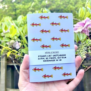 May include: A small blue sketchbook with a pattern of red and yellow fish. The cover has the text "WEIRD IS THE BEST" and "FISHIES CAT SKETCHBOOK 4X7IN, 32 PAGES, 100 GSM HANDMADE IN UK".