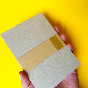 K&ouml;nnte beinhalten: Ein rechteckiges, hellbraunes Paket, umwickelt mit einem beigefarbenen Band, wird in einer Hand vor einem leuchtend gelben Hintergrund gehalten. Das Paket scheint aus Pappe oder einem &auml;hnlichen Material zu bestehen.