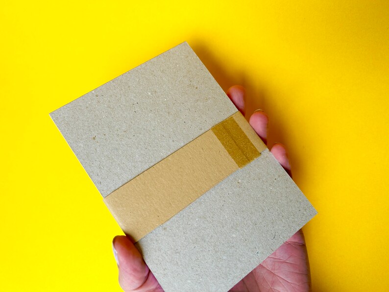 K&ouml;nnte beinhalten: Ein rechteckiges, hellbraunes Paket, umwickelt mit einem beigefarbenen Band, wird in einer Hand vor einem leuchtend gelben Hintergrund gehalten. Das Paket scheint aus Pappe oder einem &auml;hnlichen Material zu bestehen.