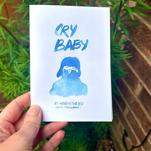 May include: A blue and white printed booklet titled "Cry Baby" by "Weird is the Best" Irena Mielczarek.