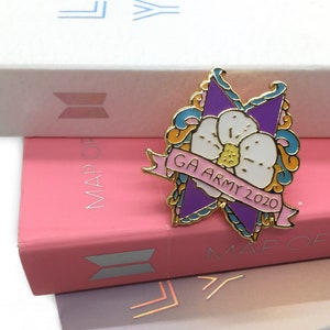 May include: A gold-colored enamel pin with a purple flower and a banner that reads "GA ARMY 2020". The flower has a white center and yellow accents. The pin is on top of a pink book with the word "MAP OF" printed on the cover.
