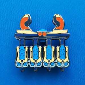 May include: A detailed enamel pin featuring a stylized amusement park ride design. The pin has a blue, yellow, and orange color scheme. The ride has four seats and a decorative top.