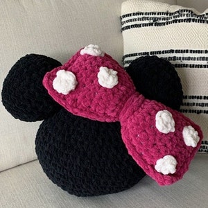 May include: A crochet Minnie Mouse pillow pattern. The pillow is black with a pink bow with white polka dots. The text "A PATTERN BY CARLI'S TREASURES" is at the bottom of the image.