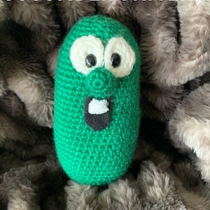 May include: A crocheted Larry the Cucumber plush toy, green with a white and black face, is sitting on a brown fuzzy blanket. The text "LARRY THE CUCUMBER PATTERN" is at the top of the image and "A PATTERN BY CARLI'S TREASURES" is at the bottom.