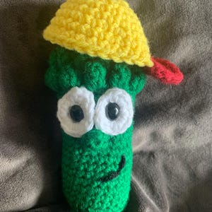 May include: A handmade crochet toy in the shape of a green character with large white-rimmed eyes and a smiling mouth. It wears a yellow hat with a red brim. The toy is made of yarn.