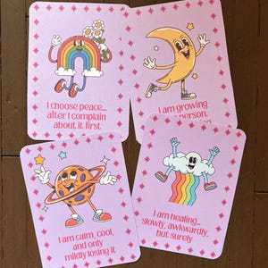 May include: Four light purple affirmation cards with cartoon illustrations of a rainbow, moon, planet, and cloud with a rainbow. Each card has a different phrase: "I choose peace...", "I am growing person.", "I am calm, cool, and only mildly losing it", and "I am healing..."