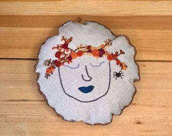 以下が含まれることがあります： 閉じた目と青い口をした茶色の刺繍が施された白い枕。顔はオレンジ、赤、茶色の花輪で囲まれています。枕の右側に小さな黒いクモが刺繍されています。