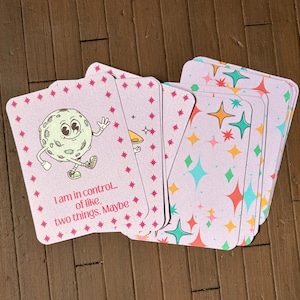 May include: A deck of tarot cards with a light pink background and a repeating star pattern. One card features a cartoon moon character with text that reads "I am in control... of like, two things. Maybe."