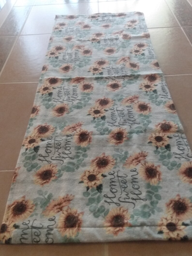 May include: A light blue table runner with a repeating pattern of sunflowers and the text "Home Sweet Home".