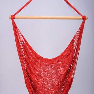 Puede incluir: Una silla hamaca roja con una barra de extensi&oacute;n de madera. La hamaca est&aacute; hecha de cuerda tejida y est&aacute; colgada de un gancho de techo.