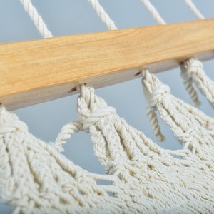 May include: Close-up of a white cotton hammock with wooden spreader bars. The hammock is knotted and has a fringe detail.