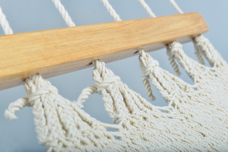 May include: Close-up of a white cotton hammock with wooden spreader bars. The hammock is knotted and has a fringe detail.