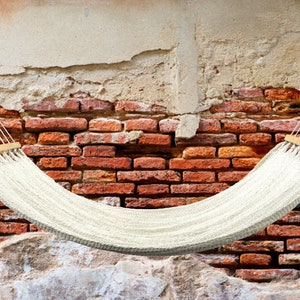 May include: A white hammock hangs between two brick walls. The hammock is made of woven rope and has wooden spreader bars.