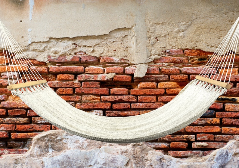 May include: A white hammock hangs between two brick walls. The hammock is made of woven rope and has wooden spreader bars.