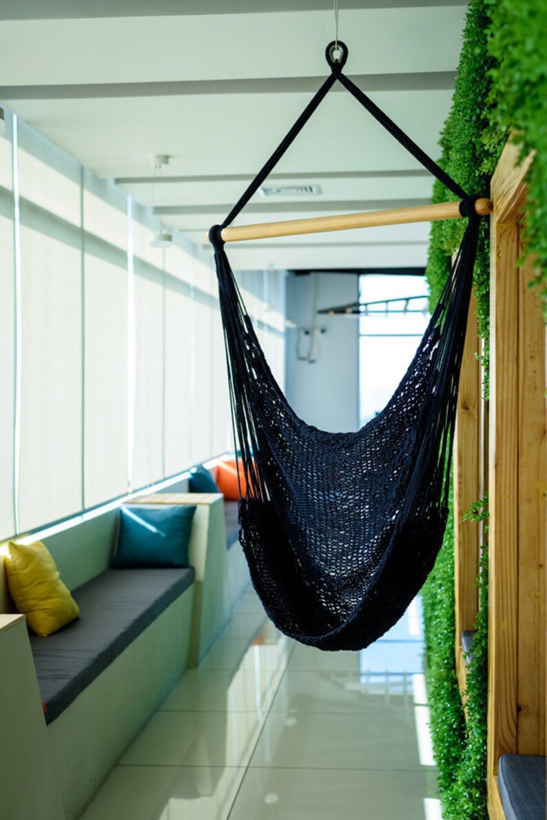 Puede incluir: Una silla hamaca negra cuelga del techo en un espacio de oficina moderno. La hamaca est&aacute; hecha de un material de cuerda tejido y tiene una barra de separaci&oacute;n de madera. La hamaca est&aacute; colgada frente a una pared con plantas verdes.