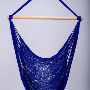 Puede incluir: Una silla hamaca azul con una barra de extensi&oacute;n de madera. La hamaca est&aacute; hecha de material de cuerda tejida y est&aacute; colgada de un lazo en la parte superior.