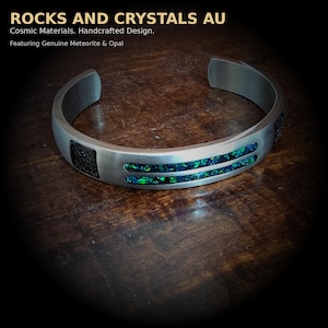 Puede incluir: Una pulsera de puño plateada con una sección negra rectangular y dos ranuras paralelas llenas de ópalo verde y azul. La pulsera está sobre una superficie de madera. El texto en la parte superior dice "ROCKS AND CRYSTALS AU".