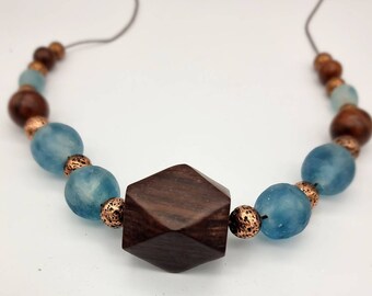 Puede incluir: Un collar hecho con cuentas de madera oscura, vidrio azul y cobre. La pieza central es una gran cuenta hexagonal de madera oscura.