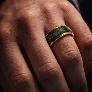 May include: Close-up of a gold-coloured ring with a dark green inlay. The ring is on a person's finger. The ring's band has a polished finish, and the green inlay has a textured appearance.