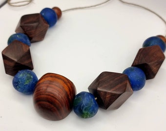 Puede incluir: Un collar hecho de cuentas de madera oscura y vidrio azul. Las cuentas de madera son facetadas y las cuentas de vidrio son redondas. El collar está encordado en un cordón marrón.