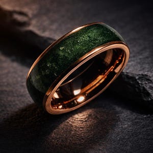 May include: A close-up of a gold-colored ring with a dark green, textured inlay. The ring has a polished finish and sits on a dark, textured surface. The ring's band is approximately 0.3 inches wide.