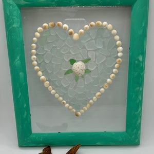 May include: A framed heart-shaped mosaic made of white sea glass and white beads with a small white seashell and green sea turtle in the center. The frame is painted a light green color.