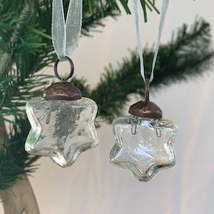 Puede incluir: Dos adornos de estrella de vidrio transparente con una cinta plateada y una parte superior de metal de color cobre. Los adornos cuelgan de un árbol de Navidad.