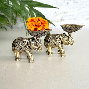 May include: Two metallic gold-toned elephant figurines, each featuring intricate embossed patterns on the body and carrying a textured, shallow bowl on its trunk. One bowl holds a vibrant orange and yellow marigold flower blossom.