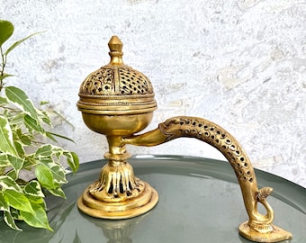 May include: A gold-colored incense burner with a decorative, perforated dome and a curved handle. The burner has a detailed base and a swan-shaped support. The design features intricate patterns and is set on a reflective, gray-green surface.
