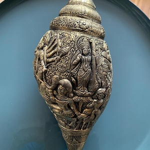 Könnte beinhalten: Eine goldfarbene Conchenschale mit aufwendigen Schnitzereien, die hinduistische Gottheiten darstellen. Die Schale ist mit floralen Mustern verziert und hat ein detailliertes Design.